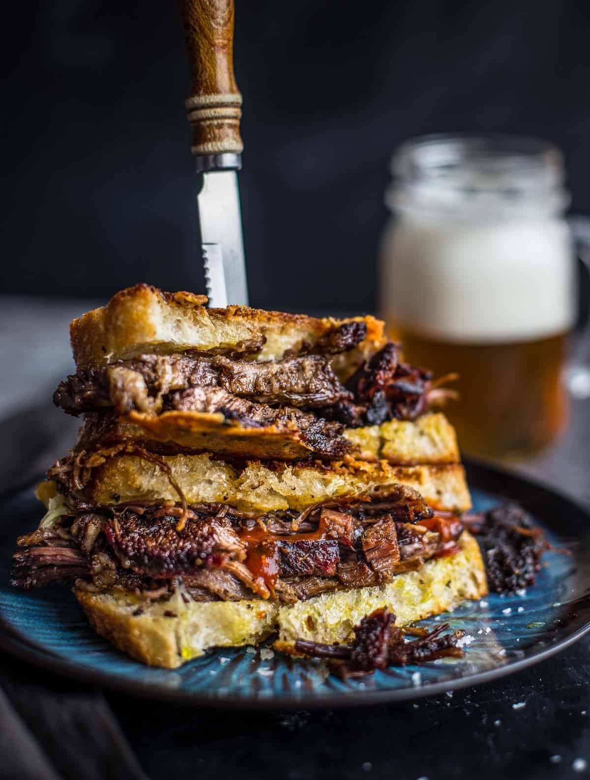 Sandwich con Brisket e Formaggio Fuso 2 Due metà di Panino al Formaggio Grigliato con Brisket, l'una sopra l'altra, con un bicchiere di birra sullo sfondo