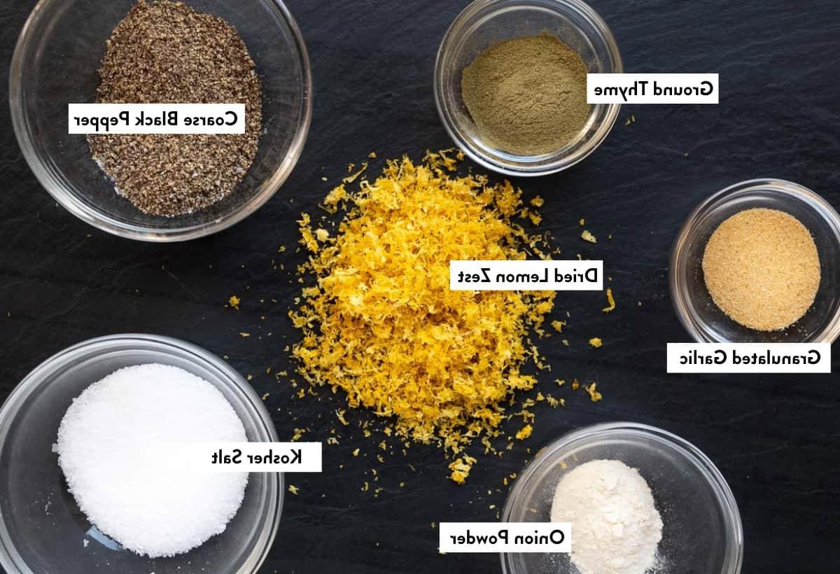 Ingredienti per il condimento fatto in casa al limone e pepe, disposti su un piatto nero ed etichettati con i loro nomi.
