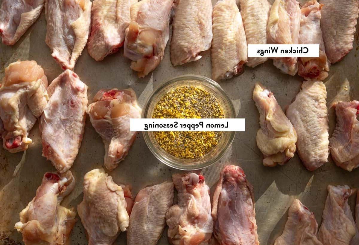 Ali di pollo crude su una teglia con una ciotola di condimento al limone e pepe fatto in casa, etichettato con i nomi degli ingredienti.