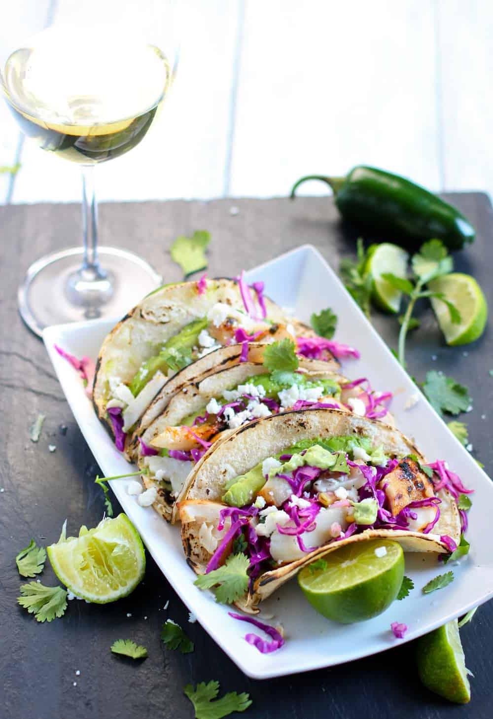 Ricetta Facile per Tacos di Pesce alla Griglia — Con Abbinamento Vino 4 Tacos di Pesce alla Griglia su un piatto bianco guarniti con lime e coriandolo, e Abbinamento Vino