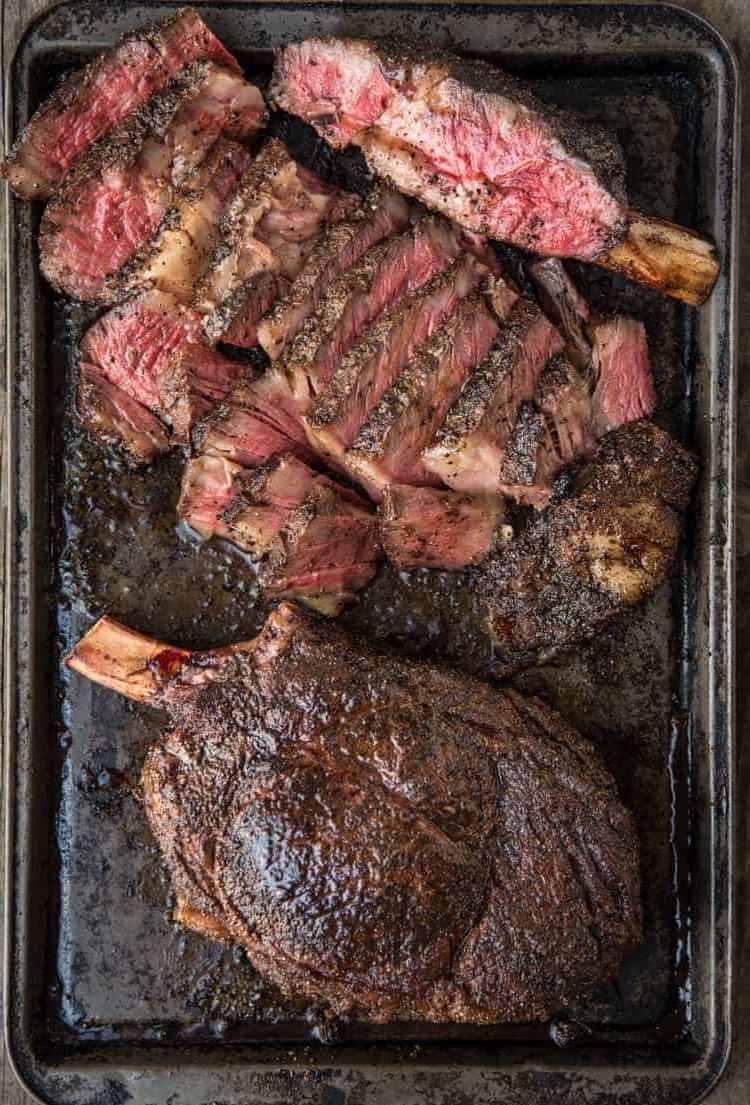 Costate Cowboy con Rub Secco al Caffè e Ancho 6 Bistecche Cowboy Ribeye alla griglia su una teglia, una intera e l'altra tagliata a strisce che mostrano il modo giusto di tagliare una grande ribeye