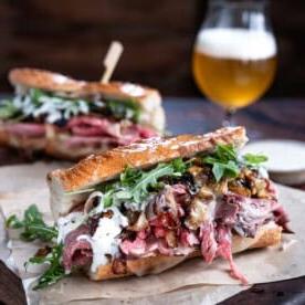 Due panini di prime rib steak con salsa al rafano su carta da forno su un tagliere con birra.