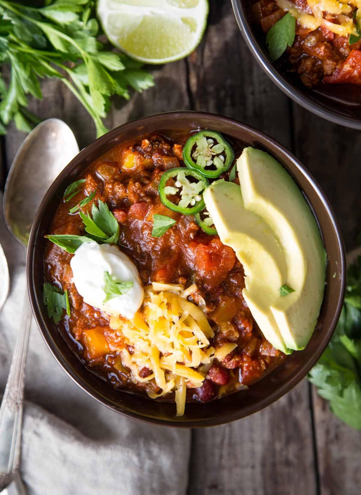 Ricetta del Chili con Carne Macinata e Peperoncini Chipotle 2 Una ciotola di chili con carne macinata guarnita con avocado, formaggio grattugiato, panna acida e fette di jalapeño.