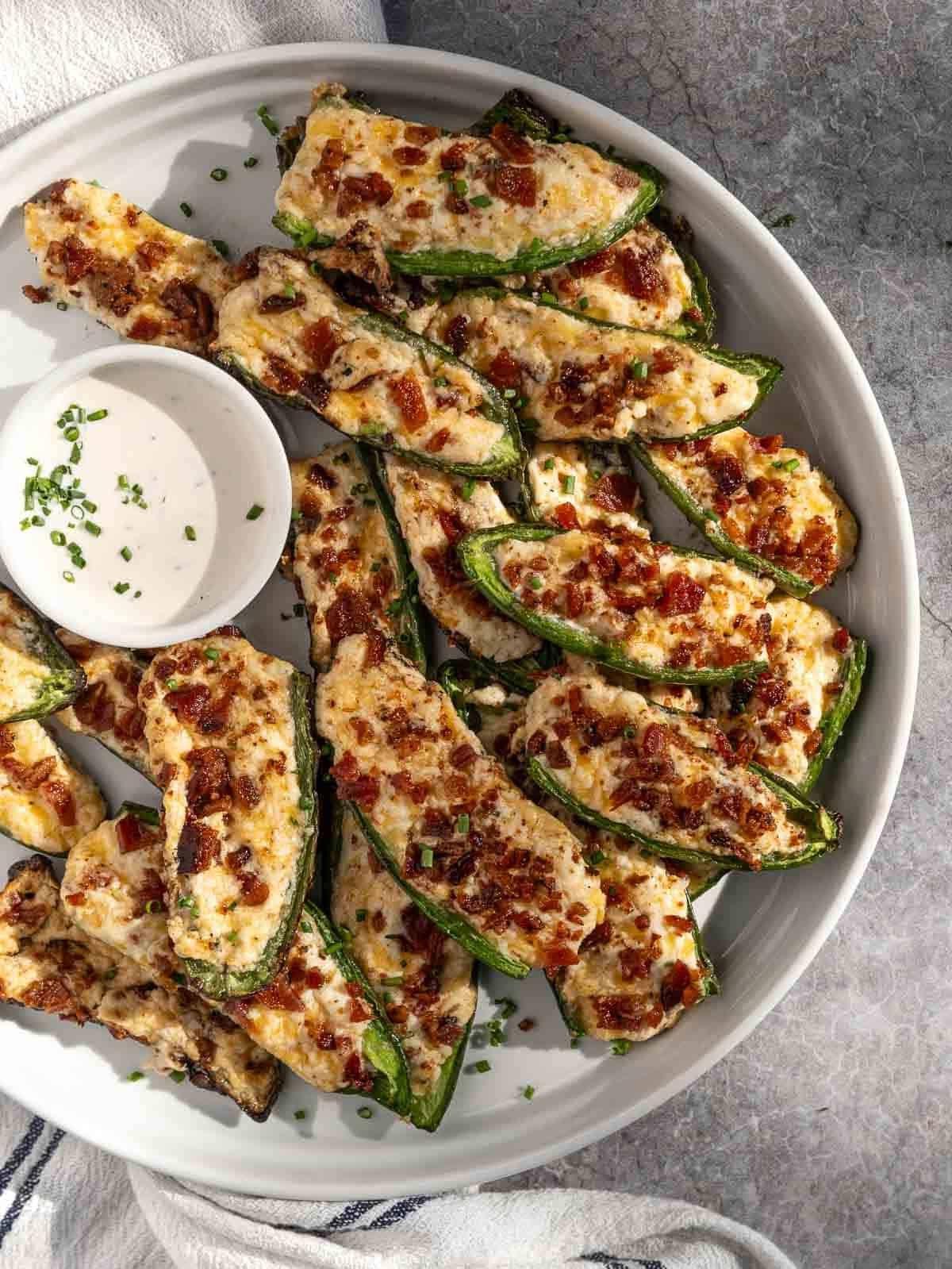 Come Preparare i Peperoncini Jalapeño Ripieni Grigliati 2 Poppers di Jalapeño Grigliati guarniti con bacon croccante su un piatto da portata con una piccola ciotola di salsa ranch.
