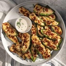 Come Preparare i Peperoncini Jalapeño Ripieni Grigliati 9 Poppers di jalapeño grigliati guarniti con bacon croccante su un piatto da portata con una piccola ciotola di salsa ranch.