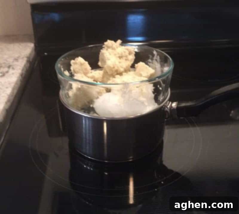 Organic raw shea butter and solid coconut oil melting gently in a glass bowl set up as a double boiler on a stovetop, creating a clear liquid mixture.