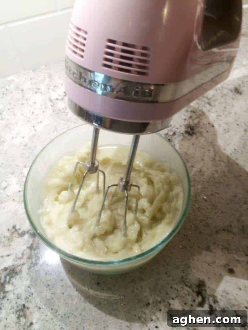 An electric hand mixer actively beating the partially solidified oil mixture in a bowl, creating a light, airy, and fluffy texture as it transforms into whipped body butter.