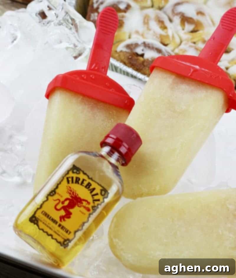 Fireball Cinnamon Roll Booze Pops 2 Fireball Whiskey Cinnamon Roll Ice Pops on tray of ice with cinnamon buns in the background and airplane-sized bottle of fireball whiskey infront of the ice pops.