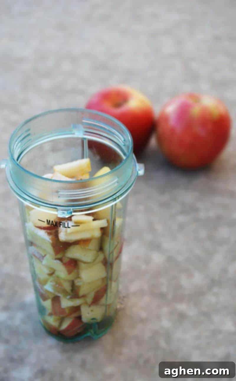 Chopped apples in a plastic cup of a blender up to max fill line. The apples are roughly chopped, with some red peel still visible, ready for blending.