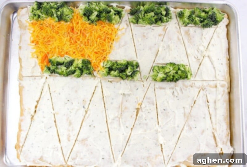 Crescent roll dough lightly scored into triangles and topped with broccoli florets, shredded cheese, and carrots, taking shape as individual veggie bars.