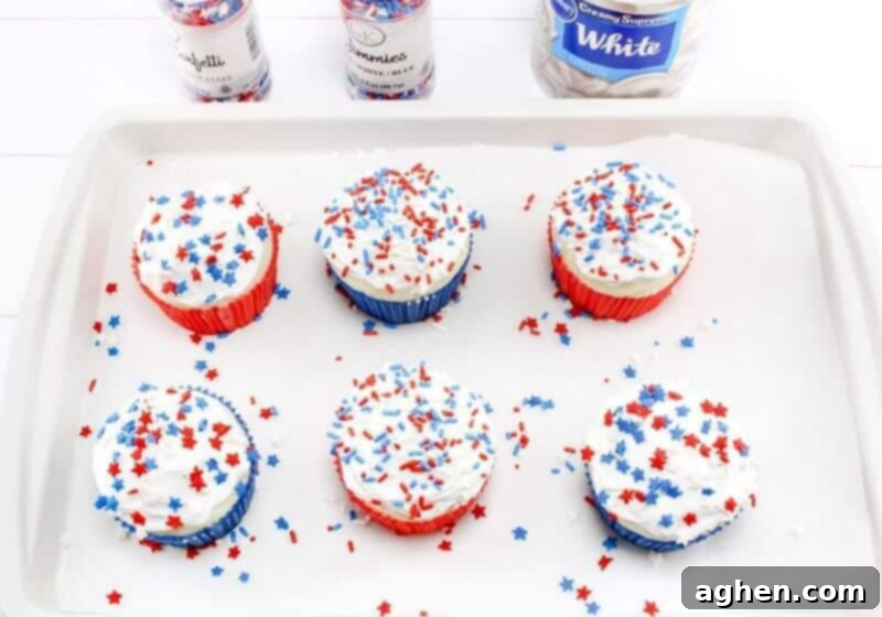 Vanilla cupcakes freshly frosted with white icing and adorned with vibrant patriotic sprinkles, ready for the firecracker toppers.