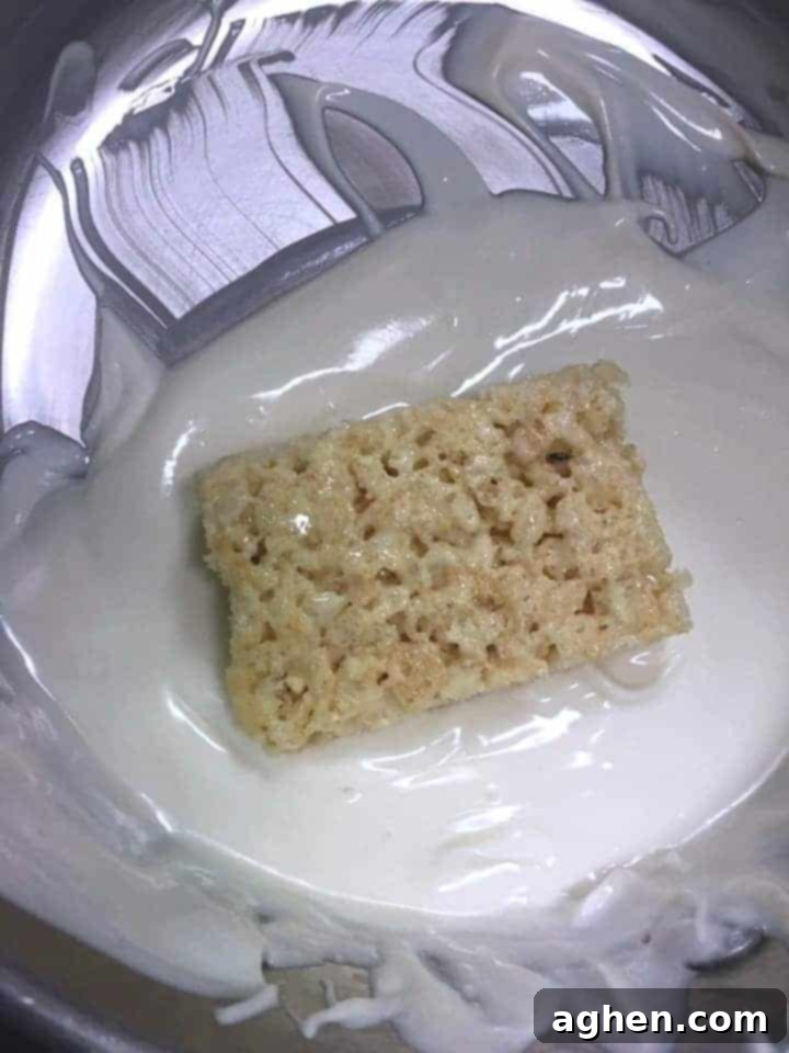 A Rice Krispie Treat being carefully dipped into a bowl of melted white chocolate, ensuring full coverage.