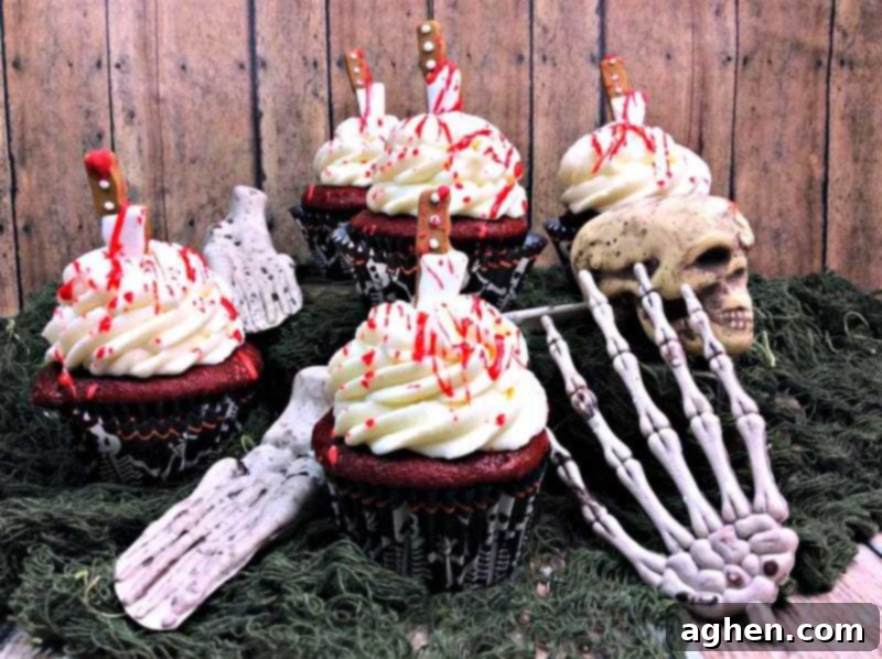 Red velvet cupcakes topped with cream cheese frosting, edible knife decorations, and edible blood splatter. Halloween skull and bone decorations surround the cupcakes on a dark surface.