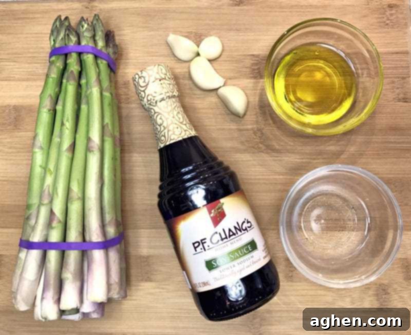 Bundle of asparagus, bottle of soy sauce, olive oil, fresh garlic cloves, and water on a counter.