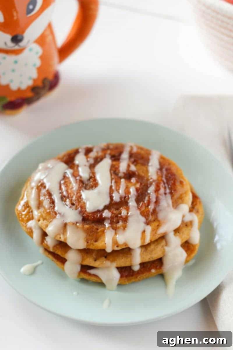 Fluffy pumpkin spice pancakes with a cinnamon swirl and a creamy white cream cheese glaze, garnished with a sprinkle of cinnamon, served on a white plate with a fork.