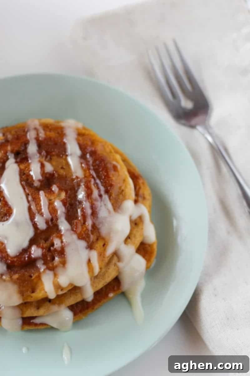 A stack of golden brown pumpkin spice pancakes with visible cinnamon swirls and generous cream cheese glaze on a white plate with a fork, ready to be enjoyed.
