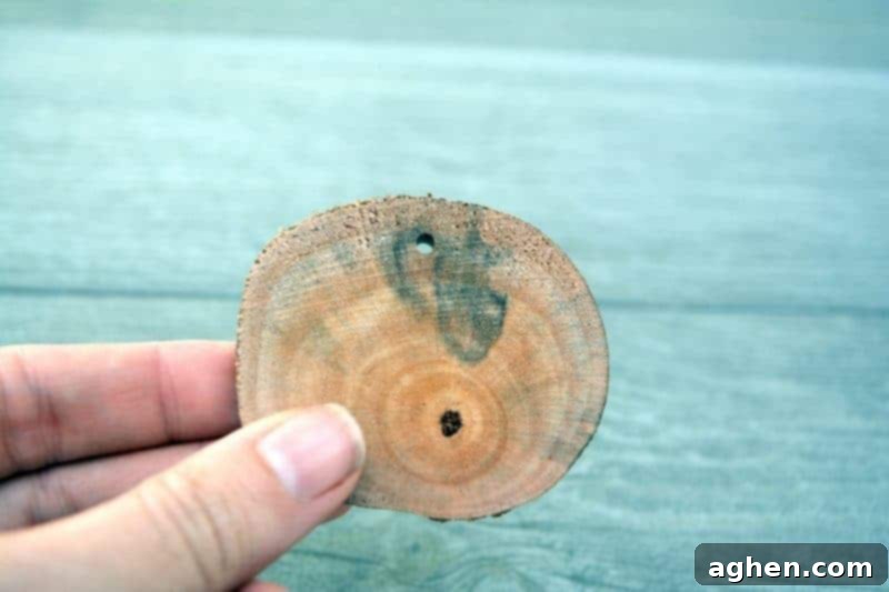 A hand drilling a small hole into the top center of a rustic wood slice, preparing it for an ornament hanger.