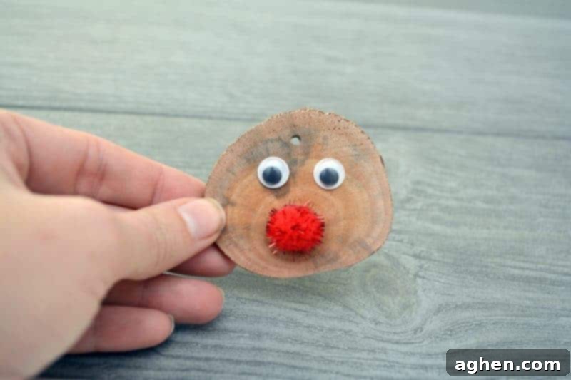 A sparkly red pom-pom glued to the lower center of a wood slice, beneath the googly eyes, forming Rudolph's red nose.