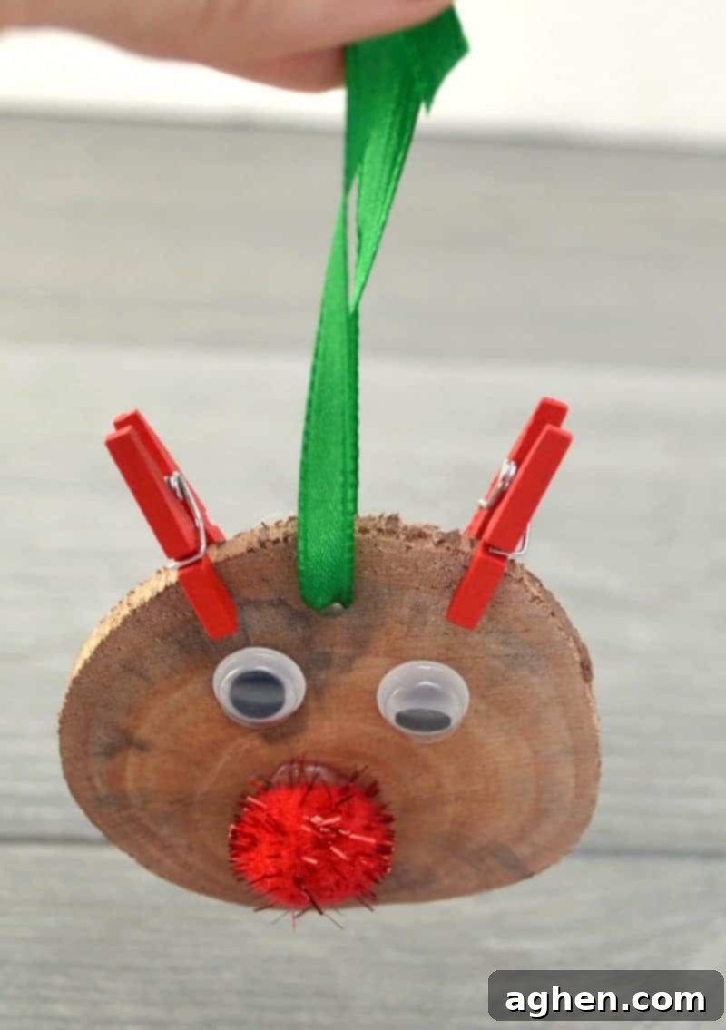 A piece of red ribbon being threaded through a pre-drilled hole at the top of a wood slice reindeer ornament.