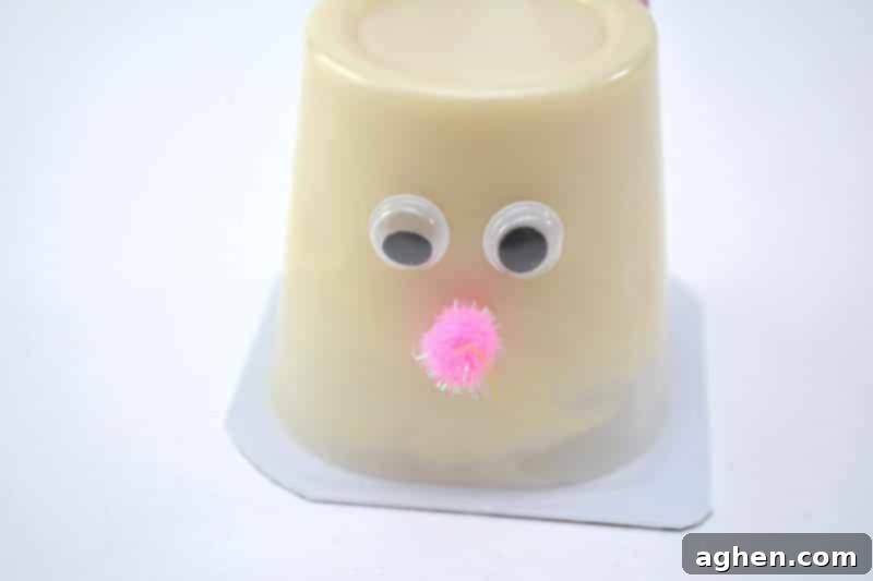 Step 7: Attach a small pink craft pom pom below the googly eyes on the front of the pudding cup to create the bunny's nose.