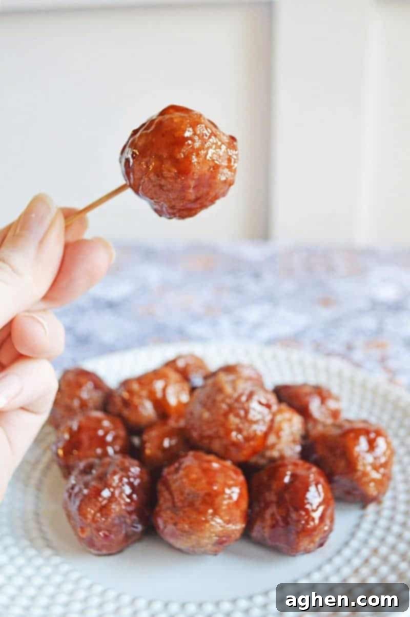 Meatballs with grape jelly and bbq sauce on a plate with a single meatball picked up with a hand holding a tooth pick.