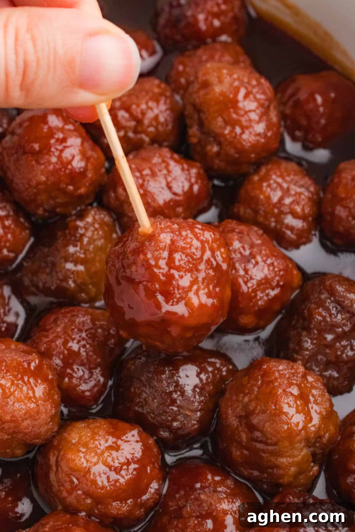 A hand lifting a grape jelly BBQ meatball from the slow cooker using a toothpick.
