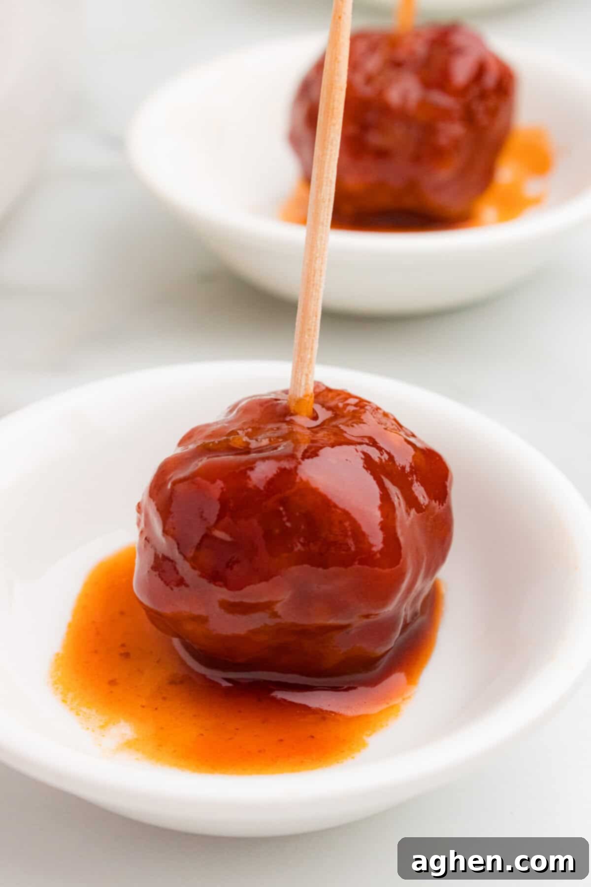 A crockpot meatball covered in thick barbecue and grape jelly sauce sits in a small white dish. A wooden toothpick is inserted in the center.