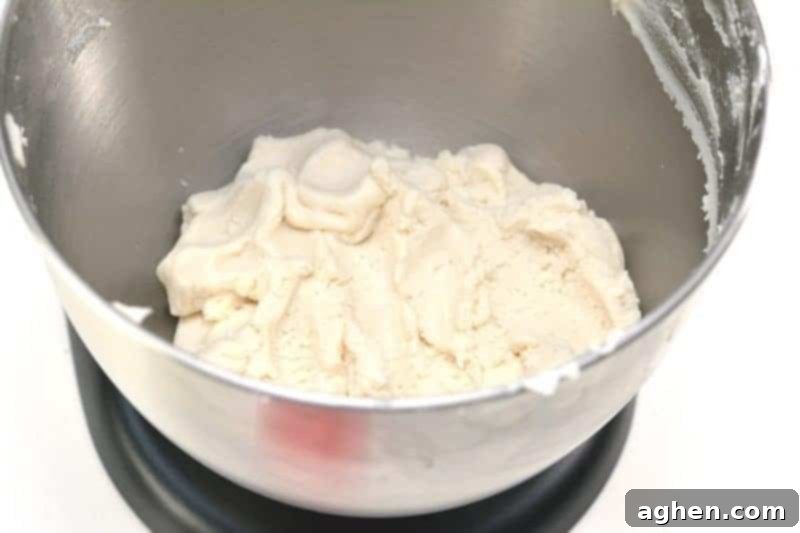The crumbly mixture of cake mix and icing in a mixer bowl, with olive oil being slowly added. This step helps achieve the desired consistency for the homemade no-cook pink princess playdough.