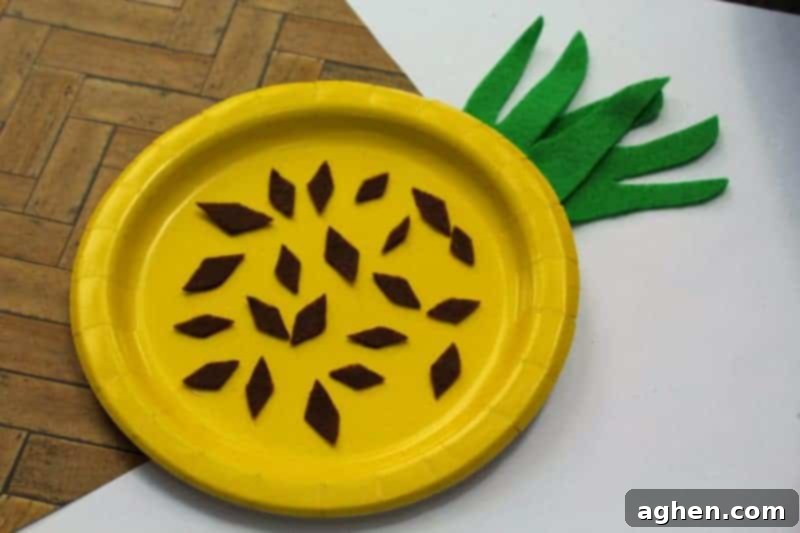 A completed paper plate pineapple craft resting on a wooden surface, showcasing its vibrant yellow color, green felt leaves, and brown felt diamond patterns, ready for display.