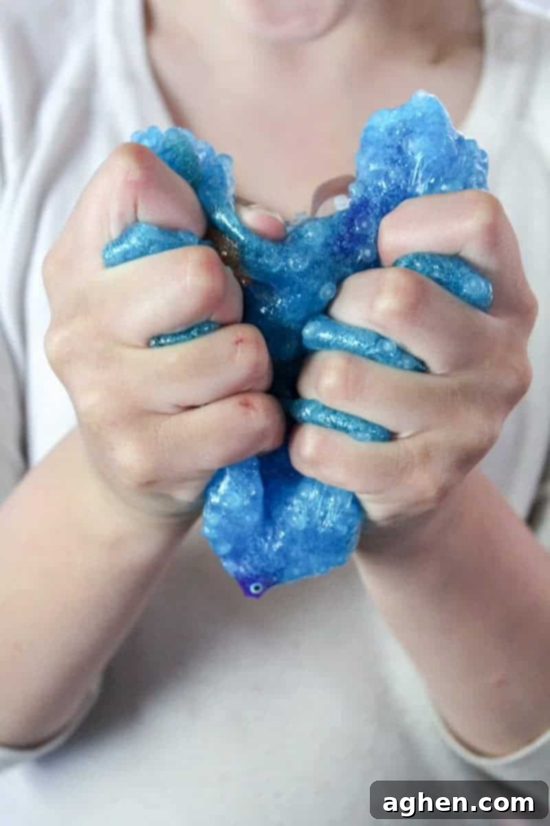Child's hands deeply engaged with blue, glittery ocean slime, stretching it to reveal embedded fishbowl beads and plastic fish, highlighting the dynamic sensory experience.