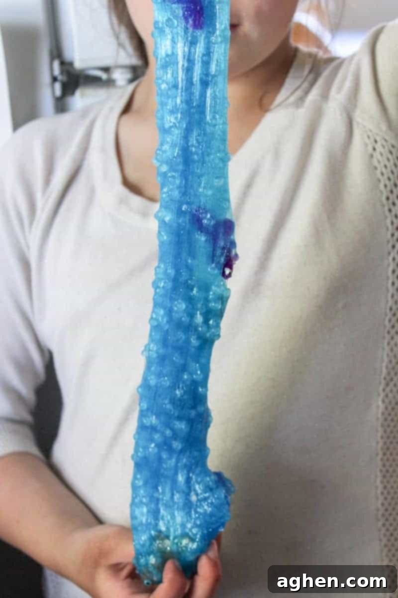 A vibrant close-up of the finished blue glitter borax-free ocean slime, beautifully stretched to display its clear, stretchy texture and the small plastic fish nestled within, ready for play.