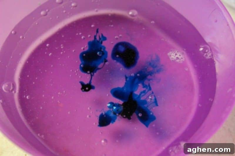 A bowl containing a clear glue and water mixture, with vibrant blue soap colorant drops being carefully added, preparing for the stirring phase.