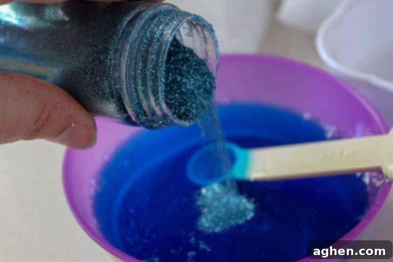 A close-up view of the blue, glittery slime mixture in a bowl, as a hand adds more sparkling blue extra fine glitter to enhance the shimmering effect.