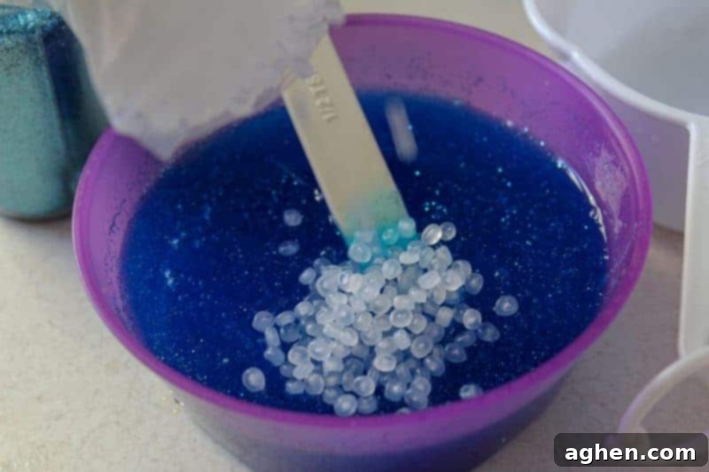 A hand gently adding clear fishbowl beads into the forming blue glitter slime in a bowl, preparing to integrate the unique texture.