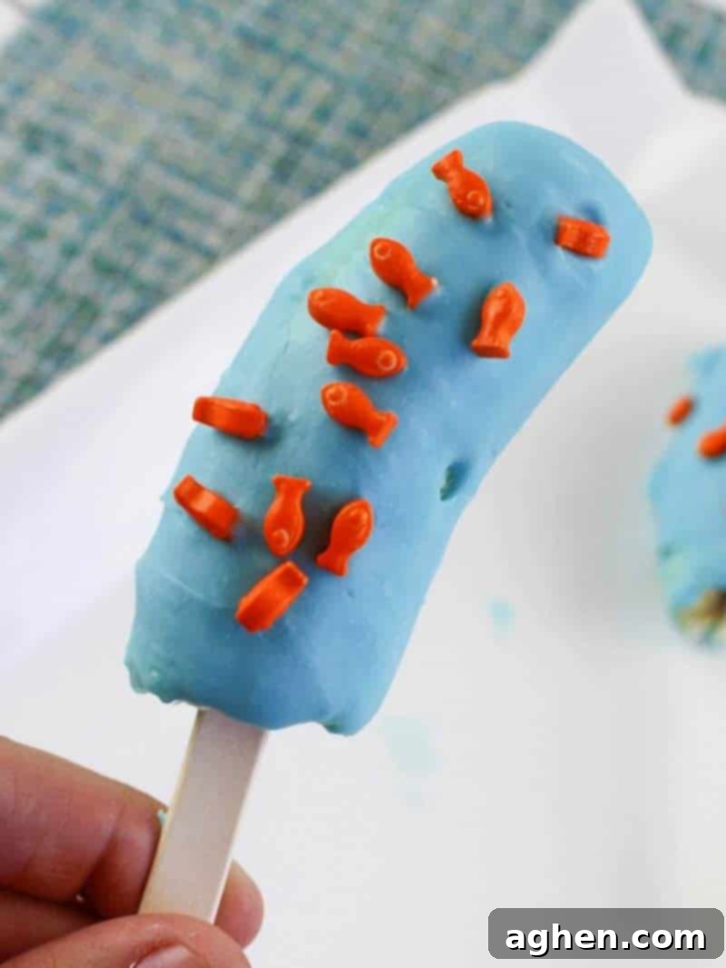 Hand holding a blue chocolate-coated banana pop with fish sprinkles on a popsicle stick.