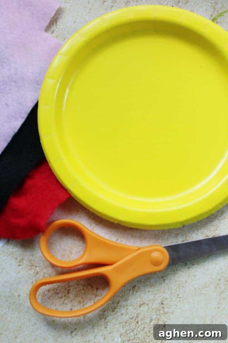 DIY Paper Plate Fish 3 Crafting supplies laid out on a white surface, including two yellow paper plates, a pair of scissors, white, black, and pink felt squares, and a bottle of craft glue.