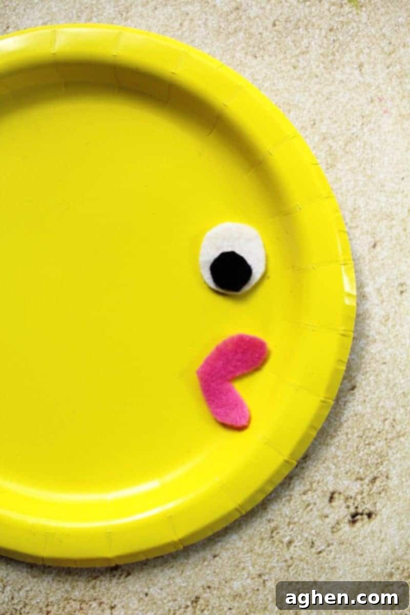 DIY Paper Plate Fish 6 A child's hands carefully gluing the pink felt lips onto a yellow paper plate, with the white and black eye pieces already attached.