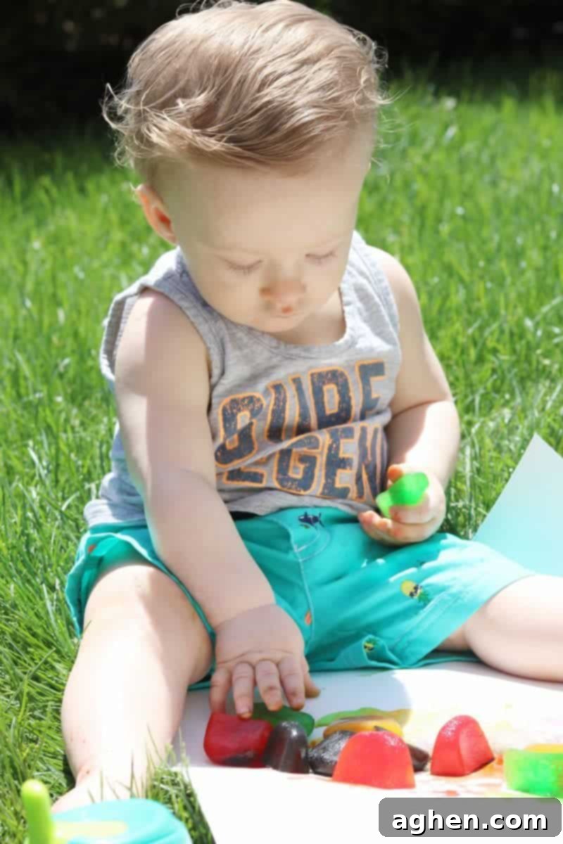 A baby's hand reaching for vibrant colored ice cubes on a white poster board, outdoors, ready for summer sensory play.