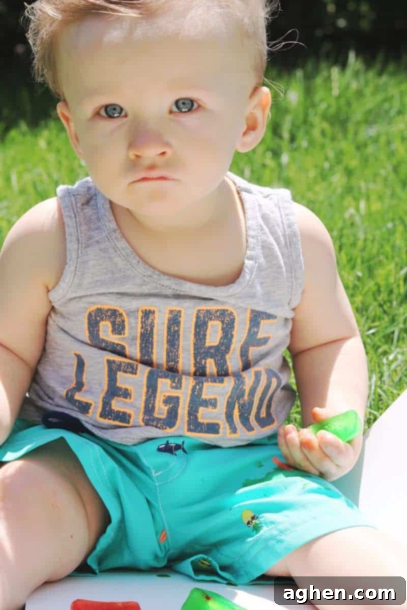 A baby sitting in the grass, surrounded by colorful melting ice cubes on a white poster board, deeply engaged in outdoor sensory play.