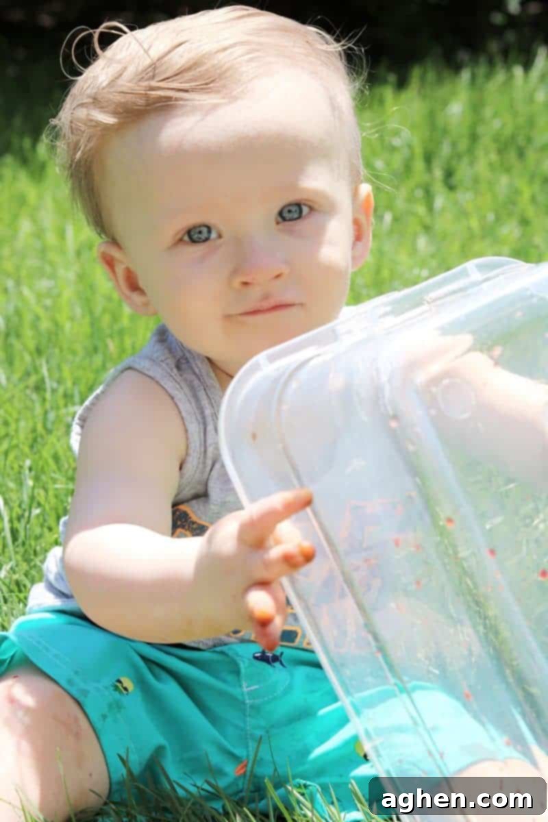 A baby gleefully tipping over a clear plastic bin, spilling numerous colorful ice cubes onto the grass during an exciting outdoor play session.