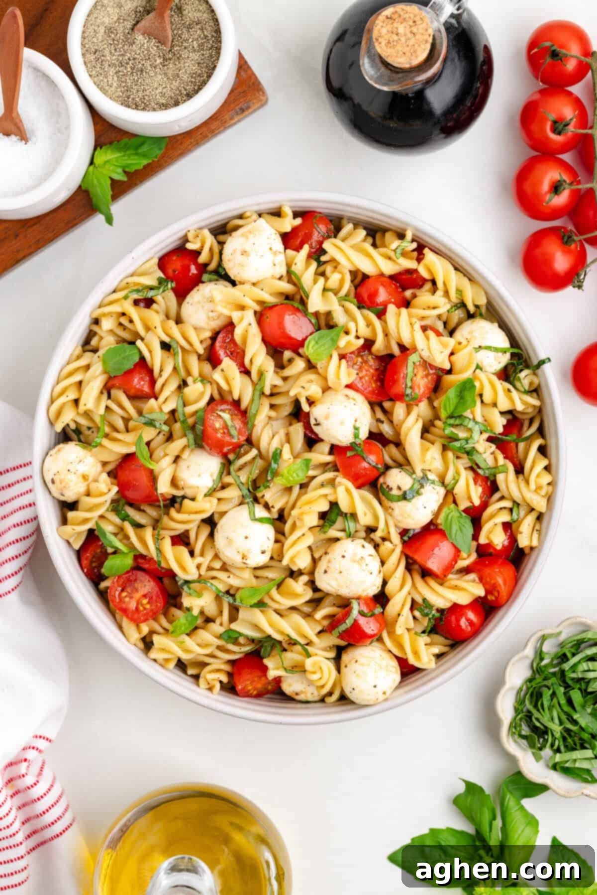 Overhead view of a large bowl of caprese pasta salad with rotini, cherry tomatoes, mozzarella balls, and basil. Around the bowl are a bottle of balsamic vinegar, salt, pepper, olive oil, fresh basil leaves, and cherry tomatoes on the vine.