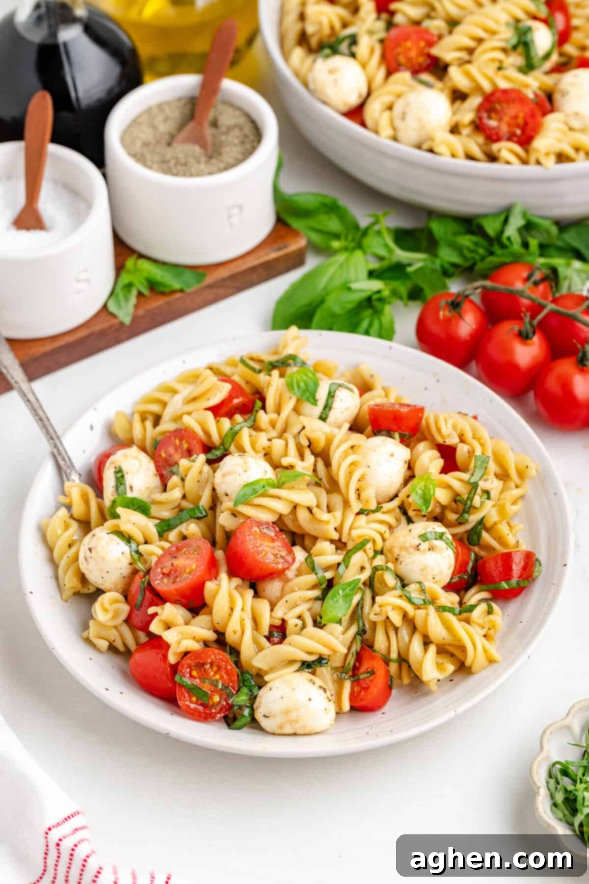 Pasta salad with basil, tomatoes and mozzarella, with ingredients in the background.