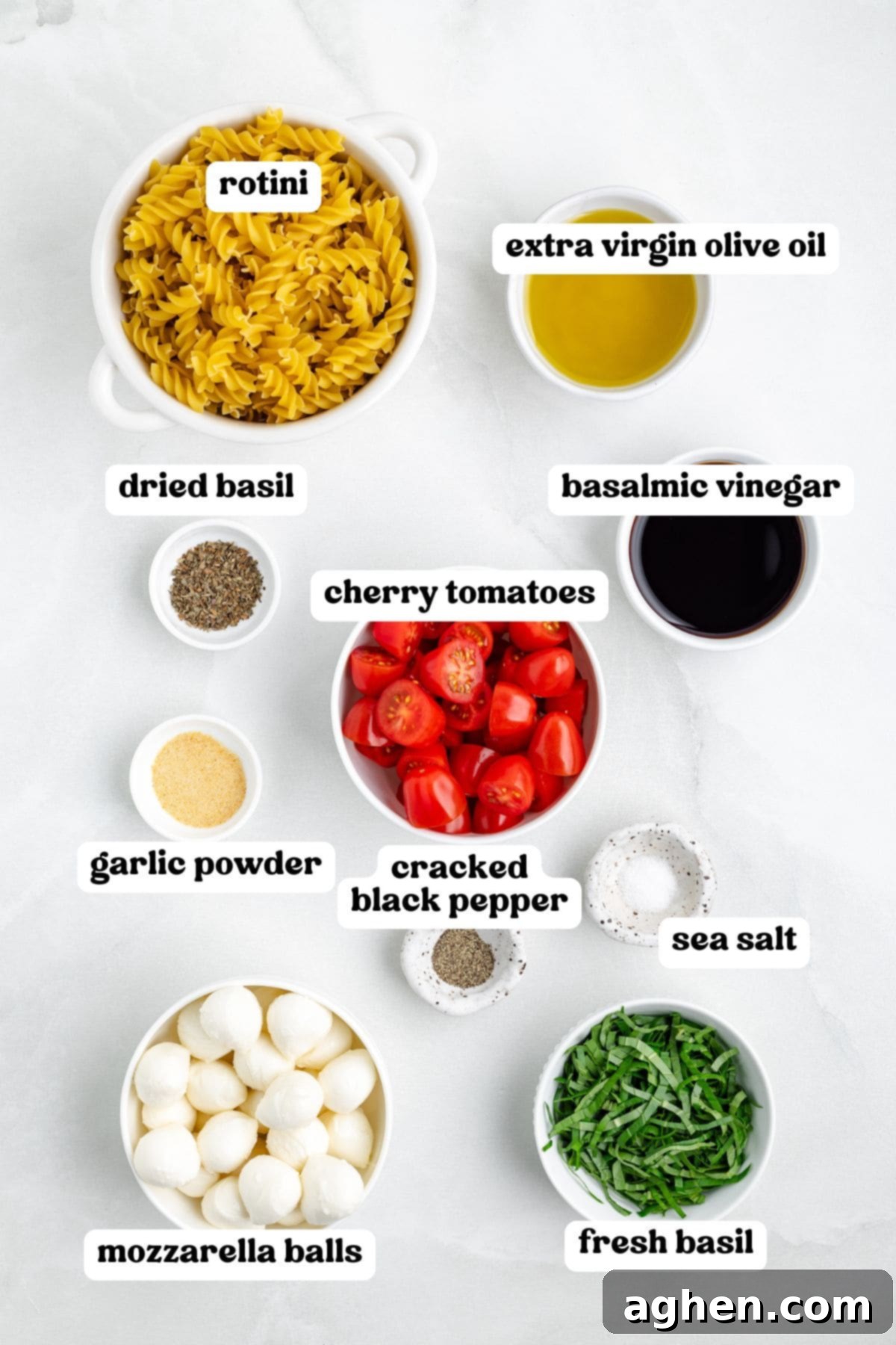 Overhead view of labeled ingredients in bowls for making Caprese pasta salad. Labels include rotini, extra virgin olive oil, balsamic vinegar, dried basil, garlic powder, cherry tomatoes, cracked black pepper, sea salt, mozzarella balls, and fresh basil.