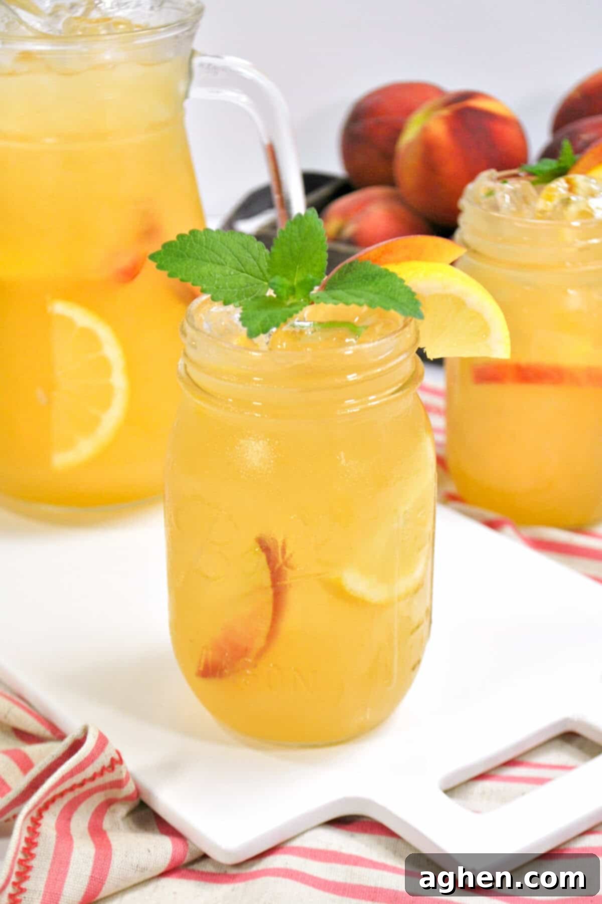 Sun Drenched Peach Lemonade 2 A mason jar of peach lemonade garnished with lemon, peach slice, and mint, with a pitcher and another jar and fresh peaches in the background.