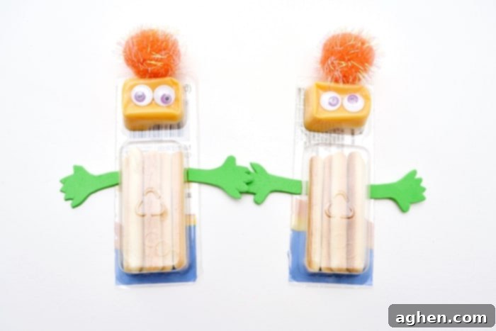 An assembled Halloween monster snack made from a packaged cheese and cracker stick, featuring green craft foam arms, googly eyes, and a pom pom.