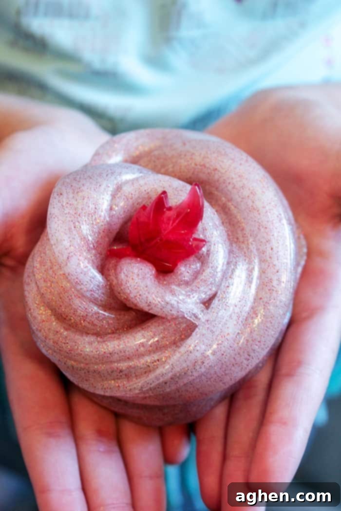 Child's hands holding cinnamon-scented fall slime with sparkling glitter and a small red plastic leaf toy on top, emphasizing sensory play.