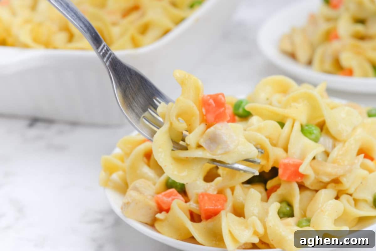 Heartwarming Chicken Noodle Bake 9 A silver fork scoops up noodles, chicken and a piece of carrot. A casserole dish and plate with a serving of chicken noodle casserole sit to the side.