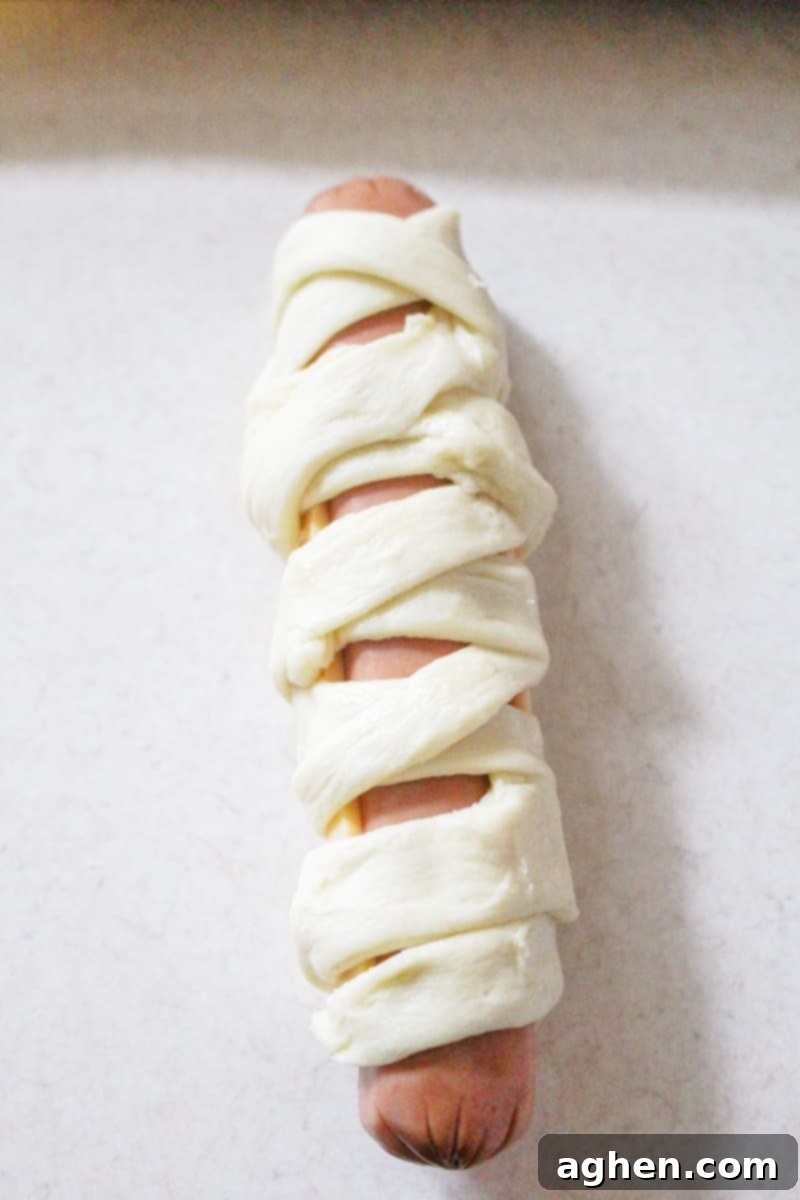 Baked mummy hot dogs, some with cheese, cooling on a baking sheet before adding the final candy eye decorations.