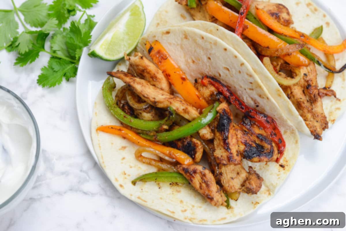 Two tortillas filled with cooked fajita chicken, peppers and onions sit on a white plate. A lime wedge sits to the side of the fajitas. Fresh cilantro leaves and a small bowl of sour cream sit to the left of the plate. 