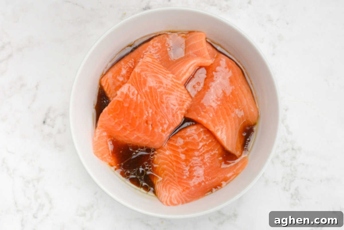 Raw salmon fillets submerged in a flavorful marinade within a clear glass bowl, covered with plastic wrap, chilling in the refrigerator.